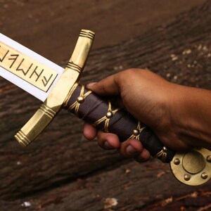 May include: A decorative sword with a gold-colored hilt and crossguard, a dark purple handle, and a blade with etched text. The sword is held in a hand against a dark wood background.