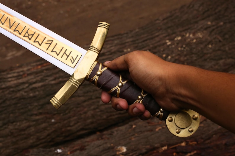 May include: A decorative sword with a gold-colored hilt and crossguard, a dark purple handle, and a blade with etched text. The sword is held in a hand against a dark wood background.