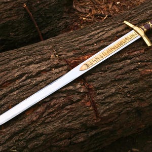 May include: A silver sword with a gold hilt and a brown and gold handle rests on a weathered log. The blade has gold lettering and a decorative emblem. The sword is a fantasy or historical item.