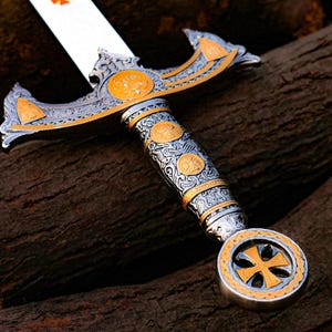 May include: A detailed close-up of a decorative sword with a silver blade and ornate handle. The handle features gold and silver accents with intricate patterns and a cross design. The sword rests on a dark wooden surface.