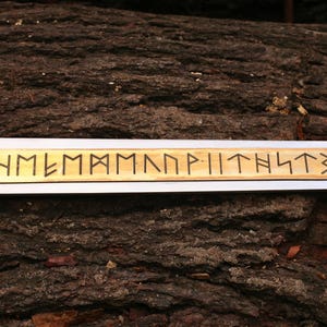 May include: A close-up of a sword with a silver blade and a gold-colored hilt. The blade has a wooden inlay with runic inscriptions. The sword rests on a textured, brown surface, possibly wood.