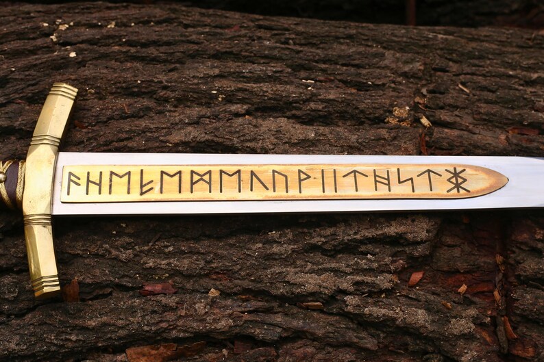 May include: A close-up of a sword with a silver blade and a gold-colored hilt. The blade has a wooden inlay with runic inscriptions. The sword rests on a textured, brown surface, possibly wood.