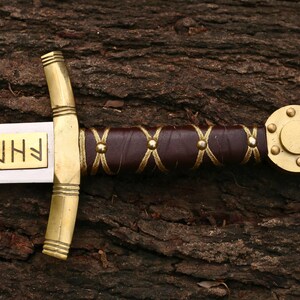 May include: A detailed view of a sword with a gold-colored hilt and accents. The handle is wrapped in brown leather with gold-colored stitching. The blade has gold lettering. The sword is resting on a textured, brown wooden surface.