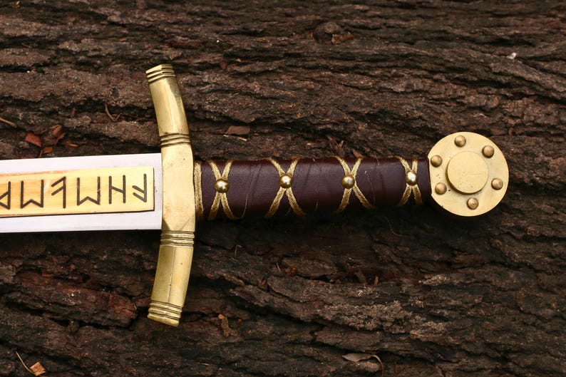 May include: A detailed view of a sword with a gold-colored hilt and accents. The handle is wrapped in brown leather with gold-colored stitching. The blade has gold lettering. The sword is resting on a textured, brown wooden surface.