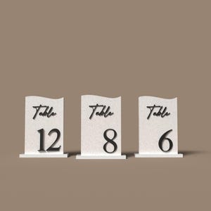 Pode incluir: Três placas brancas com números de mesa com texto preto. Cada placa tem um topo ondulado e uma base. A palavra "Table" está escrita em itálico acima de um número grande: 12, 8 e 6. As placas são para um casamento ou evento.
