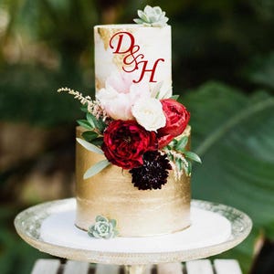 Peut inclure: Un gâteau de mariage à deux niveaux avec un glaçage doré métallique sur le niveau inférieur et un glaçage blanc sur le niveau supérieur. Le gâteau est décoré de fleurs rouges et blanches, de verdure et d'une succulente. Le niveau supérieur porte les lettres "D&H" en rouge.