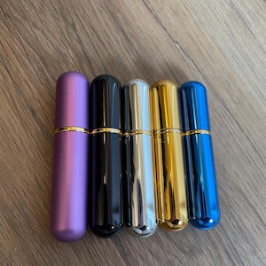 Könnte beinhalten: Eine Reihe von fünf zylindrischen Parfümzerstäubern. Die Farben sind Lila, Schwarz, Silber, Gold und Blau. Jeder hat ein goldenes Band in der Nähe der Oberseite. Der Hintergrund ist eine Holzoberfläche.