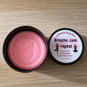 Op de afbeelding: Een ronde roze kleischijf met een vlinderopdruk in een zwart metalen blik. Het deksel bevat de tekst "Schmetterlings.MUT Breathe, calm, repeat" met yoga-iconen en de tekst "Knete mit Duft zum Entspannen, riechen, fühlen, kneten Ylang Ylang".