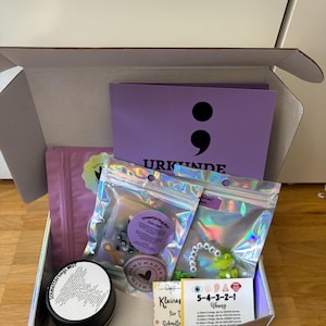 Op de afbeelding: Een kartonnen geschenkdoos met een paarse kaart met een puntkomma-symbool en het woord URKUNDE, twee iriserende zakjes met klein speelgoed en kralen, een zwart rond blik en een witte kaart met de tekst 5-4-3-2-1 Übung en iconen.