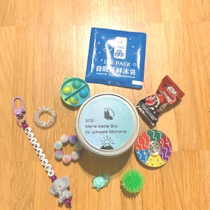 Könnte beinhalten: Eine Sammlung von Gegenständen, darunter eine runde Metalldose mit der Aufschrift SOS Meine kleine Box für schwere Momente, ein blaues Eispack, ein Schlüsselanhänger mit dem Wort MUTMACHER und Elefantenanhänger, ein Fidget-Toy, ein Bonbon und sensorische Objekte.