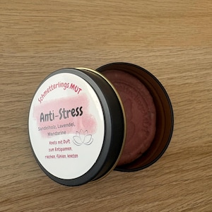 Könnte beinhalten: Eine runde schwarze Metalldose mit lilafarbener Knete. Der Deckel hat ein weißes Etikett mit rosa Aquarellakzenten, dem Text "Schmetterlings.MUT Anti-Stress Sandelholz, Lavendel, Mandarine Knete mit Duft zum Entspannen, riechen, fühlen, kneten" und einem Lotusblüten-Symbol.