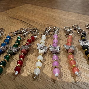 Könnte beinhalten: Eine Sammlung von Perlen-Schlüsselanhängern mit silberfarbenen Schmetterlingsanhängern, bunten, durchscheinenden Perlen, kleinen goldfarbenen Zwischenstücken und strukturierten, porösen Lavaperlen. Jeder Schlüsselanhänger hat einen silberfarbenen Karabiner.