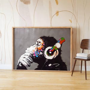 May include: A framed art print featuring a chimpanzee wearing headphones. The chimpanzee is depicted in black and white with colorful accents on the face and headphones. The print is displayed in a room with a wooden chair and a stack of books.