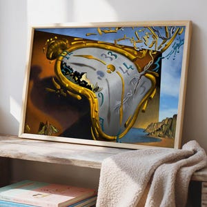 May include: A framed print of a surrealist painting featuring a melting clock with gold accents. The artwork depicts a coastal landscape with a blue sky and a brown and orange background. The print is displayed on a wooden shelf with books and a beige blanket.