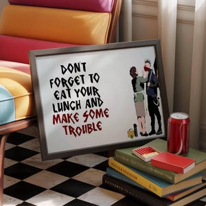 Könnte beinhalten: Ein gerahmter Druck mit dem Text "DON'T FORGET TO EAT YOUR LUNCH AND MAKE SOME TROUBLE" und einer Cartoon-Illustration. Eine rote Limonadendose und eine Streichholzschachtel liegen auf einem Bücherstapel.