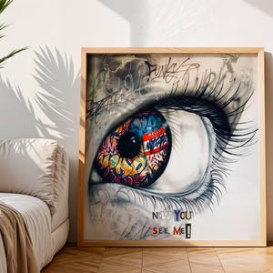 Puede incluir: Una obra de arte enmarcada que presenta un ojo estilizado con un iris vibrante de estilo grafiti. El iris está lleno de letras y diseños coloridos. La obra de arte está sobre un fondo blanco, con el texto "NOW YOU SEE ME!" en la parte inferior.