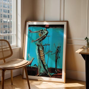 May include: A framed art print featuring a surrealist painting with a teal background. The artwork depicts a figure with outstretched arms and another figure in the background. The frame is a light brown color, and the print is leaning against a wall.