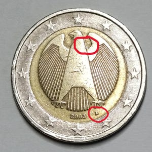 Puede incluir: Una moneda de dos euros alemana, con un detallado emblema de águila en el centro, rodeado por un anillo dorado y un anillo exterior plateado con estrellas. El año 2003 es visible debajo del águila.