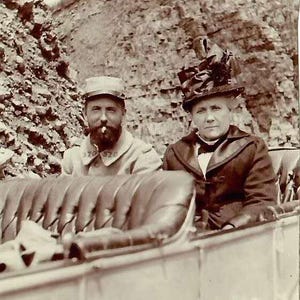 May include: A vintage sepia-toned photograph showing two individuals seated in an open-top vehicle. One person wears a military-style cap and uniform jacket with a beard, while the other wears a dark coat and a large, ornate decorative hat.
