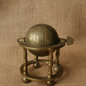 Könnte beinhalten: Eine Armillarsphäre aus Messing mit einer zentralen Kugel, die mit himmlischen Markierungen und Sternbildern graviert ist. Die Kugel wird von einem kreisförmigen Rahmen mit vier vertikalen Säulen und vier kleinen kugelförmigen Füßen getragen.