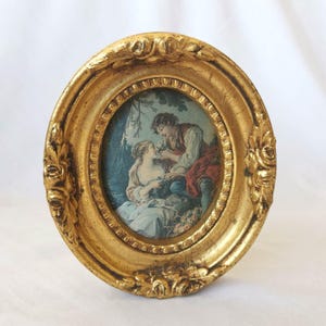 Peut inclure: Un cadre photo de forme ovale avec une finition dorée métallique et des détails en relief floral ornementaux. Le cadre contient une illustration imprimée de deux personnages en vêtements d'époque assis dans un décor naturel avec des arbres.