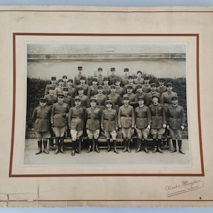 Peut inclure: Une photographie vintage en noir et blanc montée sur un carton crème avec une bordure brune, représentant un grand groupe de militaires en uniforme posant en rangs. Le texte "Clair-Guyot & Hanzikon" est imprimé dans le coin inférieur droit.