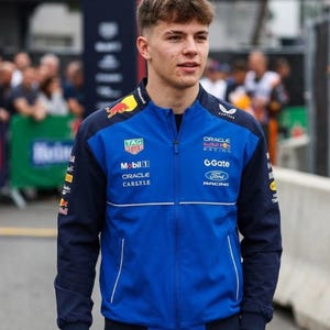 Op de afbeelding: Een persoon draagt een blauw en marineblauw racejack met meerdere sponsorlogo's, waaronder "TAG Heuer", "Oracle" en "Ford Racing". De jas heeft een ritssluiting en een opstaande kraag. De persoon staat buiten.