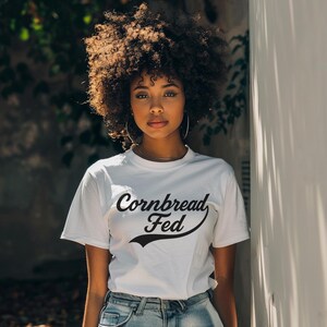 Puede incluir: Una camiseta blanca con el texto "Cornbread Fed" en escritura negra. La modelo lleva la camiseta con vaqueros azules y aros grandes. La modelo tiene el pelo rizado y está de pie frente a una pared blanca.