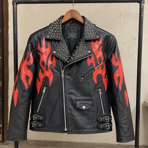 Può includere: Una giacca da moto in pelle nera con motivi a fiamma rosso brillante sulle maniche e sul petto. La giacca presenta borchie metalliche color argento sui revers, una cerniera frontale asimmetrica, due tasche con cerniera, una piccola tasca con patta e fibbie laterali regolabili.