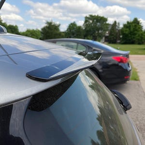 Può includere: Un'auto argentata con uno spoiler nero sul retro. Lo spoiler è montato sulla parte superiore del bagagliaio dell'auto. Lo sfondo include un cielo blu con nuvole e un'auto nera.