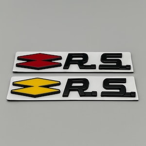 May include: Two car emblem stickers. One has a red and black diamond shape with the letters "R.S." in black. The other has a yellow and black diamond shape with the letters "R.S." in black. Both are on a white background.