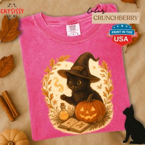 May include: A pink t-shirt with a graphic of a black cat wearing a witch hat, a jack-o'-lantern, and an open book. The shirt has the text "PRINT IN THE USA" and "CRUNCHBERRY". The brand name "GATSISSY STORE" is also visible.