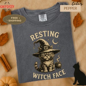 May include: A pepper-colored t-shirt featuring a grumpy cat wearing a witch hat. The text on the shirt reads "RESTING WITCH FACE". The design includes a crescent moon and a cauldron. The shirt is from the Catsissy Store.