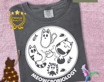 May include: Pepper-colored t-shirt with a white graphic of cartoon cats and the text "MEOWCROBIOLOGY." The shirt features a crew neck and a Comfort Colors tag. A gold "Premium Quality" emblem is visible.