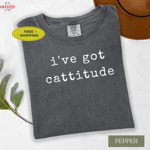 May include: A dark gray t-shirt with the phrase "i've got cattitude" in white lettering. The shirt has a crew neck and short sleeves. A brown hat is partially visible in the background. The shirt is labeled "Pepper".