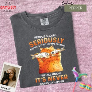 May include: Pepper-colored t-shirt with a graphic of an orange cat holding fish, and the text "PEOPLE SHOULD SERIOUSLY STOP EXPECTING NORMAL FROM ME. WE ALL KNOW IT'S NEVER GOING TO HAPPEN." The shirt has a crew neck and short sleeves.