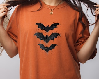 May include: A burnt orange short-sleeve t-shirt featuring a graphic print of three black bats stacked vertically in the center. The bat illustration includes subtle paint splatter details and a distressed texture, set against the solid fabric.