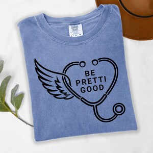 May include: A blue t-shirt with a black graphic of a stethoscope forming a heart shape with wings and the words "BE PRETTI GOOD". The shirt is folded and has a Comfort Colors tag.