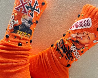 May include: A pair of orange socks with various embellishments. One sock features a graphic of a person and the word "KISSSSS". The other sock has a graphic of a person, the word "REEZY", and the letter "X". Both socks have black and gold accents.