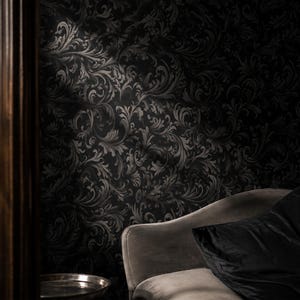 Könnte beinhalten: Eine dunkle Tapete mit einem sich wiederholenden schwarzen und anthrazitfarbenen Damastmuster mit komplexen, wirbelnden Akanthusblattmotiven. Die Wand wird teilweise beleuchtet, was die Textur des verzierten Blumendesigns hervorhebt.