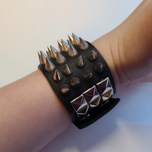Może przedstawiać: Czarna skórzana bransoletka ze srebrnymi stożkowymi ćwiekami i kwadratowymi metalowymi akcentami. Bransoletka ma szeroki styl mankietu z wieloma rzędami ćwieków. Metalowe akcenty mają geometryczny wzór.