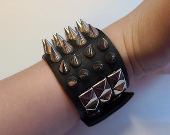 May include: Black leather bracelet with silver-colored cone studs and square metal accents. The bracelet is a wide cuff style, with multiple rows of studs. The metal accents have a geometric design.