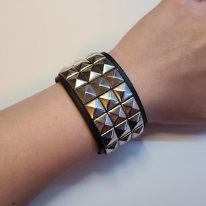 May include: A wide black leather bracelet with three rows of large, silver pyramid studs. The bracelet is worn on a wrist and has a classic punk rock aesthetic. The studs are shiny and reflect light.