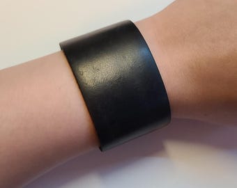 May include: A wide, black leather cuff bracelet. The bracelet is a solid band of black leather, worn on the wrist. The leather has a slightly textured appearance.