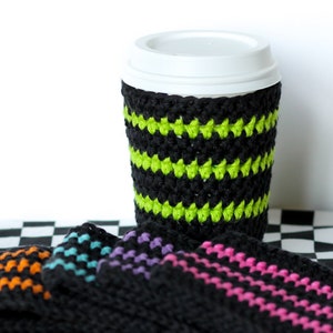 Pode incluir: Quatro capas de crochê para caneca de café em preto com listras coloridas. As capas estão dispostas em uma superfície quadriculada preta e branca. Uma das capas está em uma caneca de café branca.