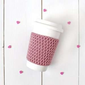 Pode incluir: Uma caneca de café para viagem branca com um aconchegante de crochê rosa. A caneca está cercada por corações rosas sobre um fundo de madeira branca.