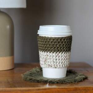 May include: A white coffee cup with a green and white crocheted cosy. The cosy has a textured pattern and is sitting on a brown woven coaster.