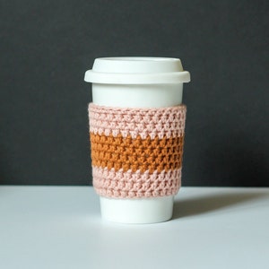 May include: A white ceramic travel mug with a white lid and a crocheted cozy. The cozy is made of pink and orange yarn, creating horizontal stripes. The mug is designed to keep beverages warm.