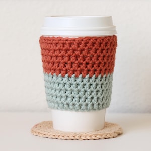 Pode incluir: Um aconchegante de xícara de café de crochê em cores ferrugem e verde-sálvia. O aconchegante é feito com ponto duplo de crochê e é projetado para caber em uma xícara de café de tamanho padrão.