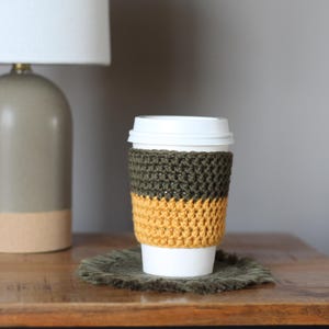 Può includere: Un copri-tazza da caffè all'uncinetto con uno schema di colori verde e giallo. Il copri-tazza è su una superficie di legno marrone.